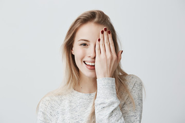 Wall Mural - Happy smiling female with attractive appearance and blonde hair wearing loose sweater showing her broad smile having good mood closing her eye with hand, enoying to pose at camera. Happiness and joy