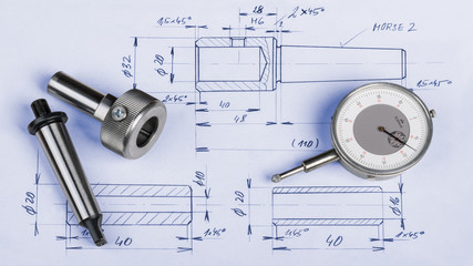 Metal engineering components, gauge and technical drawing. Steel machining jigs and dial gauge micrometer laid on the manufacturing documentation.