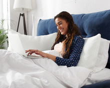 Happy casual beautiful woman laughing during work on a laptop sitting on the bed in the house