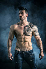 handsome shirtless muscular man with jeans, standing, on dark smoky background in studio shot