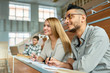 © Seventyfour - Multi-ethnic group of students sitting at desk in lecture hall of modern college and smiling happily, focus on young Middle-Eastern man wearing glasses, copy space