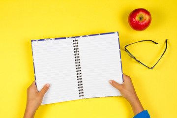 Kid hand with open notebook, glasses and red apple on yellow background