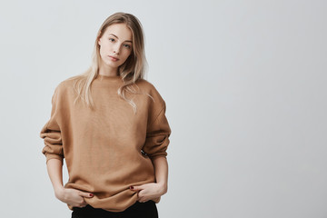 Wall Mural - Cute pretty beautiful woman wears loose sweater and black trousers, holds her hands in pocket, looks with appeal at camera. Pleasant-looking girl posing against gray background