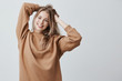 © Cookie Studio - Beautiful pretty charming young blonde woman in loose sweater smiling happily, having fun indoors, playing with long straight hair. Pretty girl looking at camera with joyful smile, rejoicing life