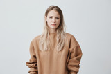 Fototapeta  - Dissatisfied blonde young woman wearing long sleeved sweater frowning face, looking angrily at camera, being displeased with her husband`s words. Negative emotions and reaction