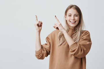 Sticker - Advertising concept. Excited cheerful european woman with long blonde hair, wearing casual clothes and smiling happily, pointing index fingers upwards, motivating and attracting customers.