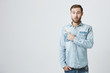 Amazed shocked male model with beard, wearing denim shirt, looking in surprisement at camera with opened mouth, pointing with forefinger at copy space for your text or advertisement