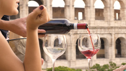 Two happy young woman tourists sitting at the table of a bar restaurant in front of the Colosseum in Rome drink and toast with a glass of italian red wine. Stylish colorful dress on a summer day at