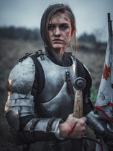 Portrait Of Girl In Image Of Jeanne D'Arc In Armor With Flag And Sword In Her Hands On Meadow.