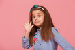 Portrait of funny female kid having long auburn hair looking on camera giving high five meaning hi or bye with hand over pink background