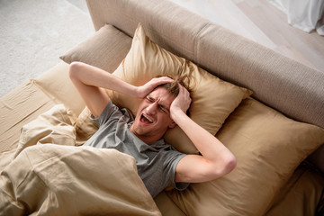 Feeling exhausted. Terribly tired out guy lying in bed. He is screaming and holding his head. He is having a headache in the morning in bedroom