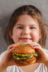 The little girl is eating a healthy baked sweet potato burger with a whole grains bun, guacamole, vegan mayonnaise and vegetables. Child vegan concept, gray background.