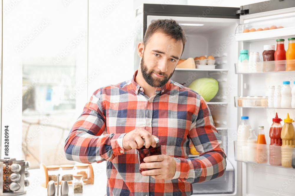 Handsome man opening jar near refrigerator in kitchen