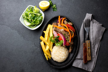 Wall Mural - Peruvian dish Lomo saltado - beef tenderloin with purple onion, yellow chili, tomatoes served on black plate, gray slate background, top view