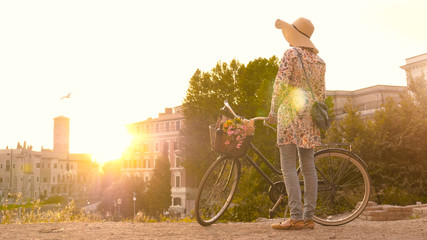 Young woman tourist with bycicle takes pictures of the colosseum in rome at sunset with smartphone. Beautiful stylish dress with large hat, flowers and bread in basket.