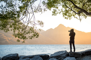 Wall Mural - Young male photographer taking photo under the tree at Lake Wakatipu during golden hour sunset in Queenstown, South island, New Zealand, travel and landscape photography concepts