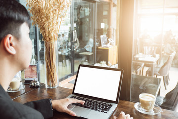 Mockup image of business man using and typing on laptop with blank white screen and coffee cup on glass table in modern loft cafe, Soft focus on vintage wooden table.