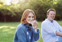 Outdoor Portrait Of Smiling Woman Drinking From Glass With Laughing Man In Background