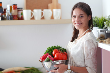 Wall Mural - Smiling young woman holding vegetables standing in kitchen. Smiling young woman