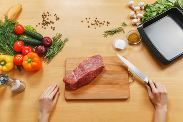 Wall Mural - Raw meat. Fresh beef tenderloin on cutting board on wooden table with different vegetables, spices, mushrooms, baking tray, knife. Top view flatlay. Copy space for advertisement. Woman cuts with knife