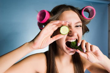 Wall Mural - funny young girl holding a cucumber for moisturizing the skin of the face, on the head of curlers