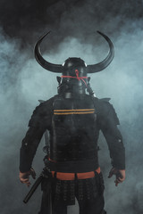 back view of samurai in traditional armor and horned helmet on dark background with smoke