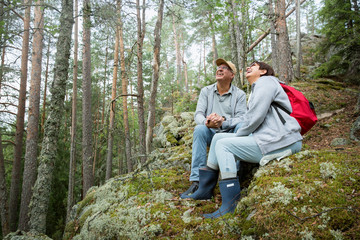 Loving senior couple hiking, sitting on the top of rock in forest, exploring. Active Mature man and woman hugging and Happily smiling. Scenic view. Healthy lifestyle. Finland.