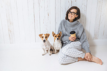 Smiling cheerful young female wears warm woolen sweater, square eyewear, drinks hot beverage, relaxes on floor and her two favourite pets, look directly into camera. People and animals conecpt