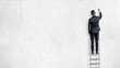 © gearstd - A businessman stands on a step ladder in a back view and draws on an empty white wall.