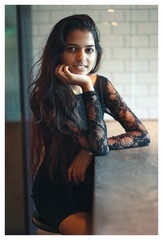 Wall Mural - Portrait of young Indian Asian woman sitting at a cafe table, resting her face on her hand and smiling at the camera.  