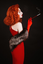 Red-haired Woman With Curly Hair In Red Dress And Long Gloves Smoke On Black Background. Girl Is Dressed In Retro Style With Natural Fur And The Mouthpiece With A Cigarette