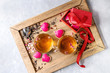 Love Valentines day greeting card with two glasses of hot tea, rose buds, heart shape homemade cookies as gift, pink sugar, tea strainer, red ribbon on wooden tray over gray background. Top view