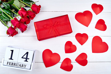 Red gift box with ribbon bow, hearts, bouquet of red roses and wooden block calendar february 14 on white background. Greeting card mockup for Valentines Day. Love concept. Top view, flat lay