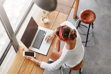 Prosperous young talented female editor checks work of employee on notebook, holds smart phone as waits for important call, sits at coffee shop, going to meet with fashion magazine director.