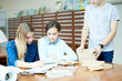 © Seventyfour - Group of students gathered together in spacious library and working on ambitious university project, unrecognizable groupmate standing at wooden table and leaning on pile of books