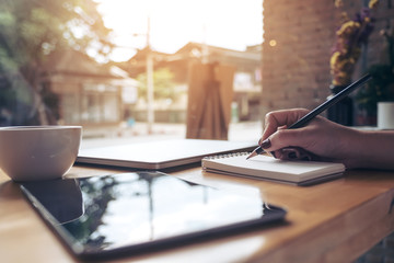 Closeup image of woman's hand writing on a blank notebook with laptop , tablet and coffee cup on wooden table background