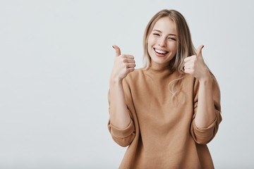 Wall Mural - Portrait of fair-haired beautiful female student or customer with broad smile, looking at the camera with happy expression, showing thumbs-up with both hands, achieving study goals. Body language