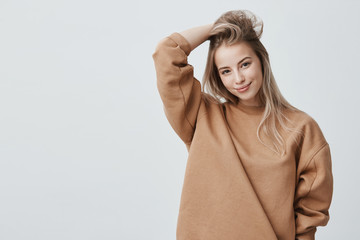 Wall Mural - Young attractive woman wearing stylish long-sleeved sweatshirt looking with her dark appealing eyes at camera, smiling gently. Blonde caucasian female student posing against gray studio wall