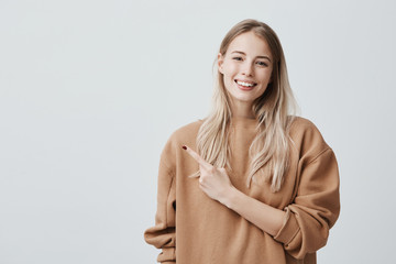 Wall Mural - Check this out! Cropped studio shot of attractive good-looking smiling young woman in casual clothes pointing with index finger up having pleased look, with cheerful and happy expression of face.