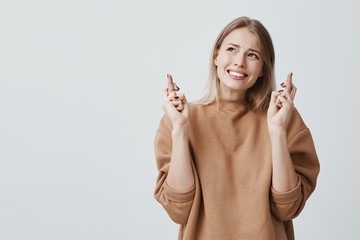 Wall Mural - Indoor shot of blonde student girl dressed in beige sweater, crosses fingers and has great hope for better, poses against gray background. Young female student wishes fortune before exam