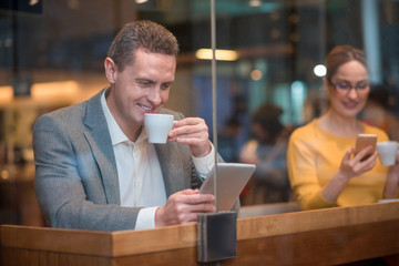 Poster - Portrait of cheerful male watching at electronic tablet. Happy girl typing in mobile. They tasting mugs of tea while situating at desk. Relax concept