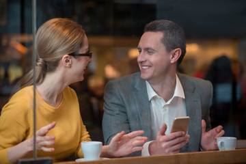Poster - Outgoing woman and happy male speaking while situating at desk in restaurant. He holding phone in arm. Leisure and technology concept
