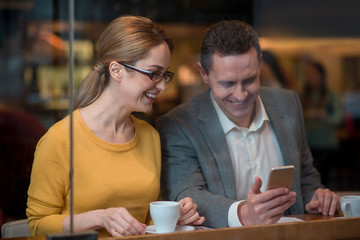 Poster - Cheerful female and smiling male looking at mobile while tasting mug of beverage. They sitting at table in cafe. Relax concept
