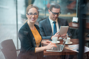 Poster - Portrait of cheerful businesswoman typing in laptop while sitting at desk. Calm colleague looking at electronic tablet. Occupation concept