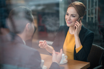 Poster - Portrait of beaming businesswoman talking by phone while situating at table with colleague. He turning back to camera. Communication and relax concept