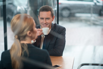 Poster - Portrait of beaming male drinking cup of coffee while telling with female partner. he situating at desk in cafe. Break concept
