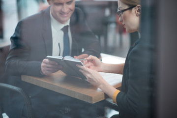 Poster - Smiling businessman and calm female watching at appointment book. They sitting at desk indoor. Career and discussion concept