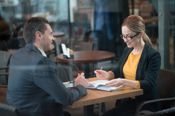 Poster - Portrait of outgoing young woman writing in appointment book while sitting at desk. She speaking with smiling comrade. Communication and job concept