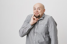 Portrait Of Bald Bearded Man With Funny Expression Holding His Pinky Near Mouth Over White Background. Guy Is Sure Up To Something, Thinking About Something Evil And Bad In His Mind.