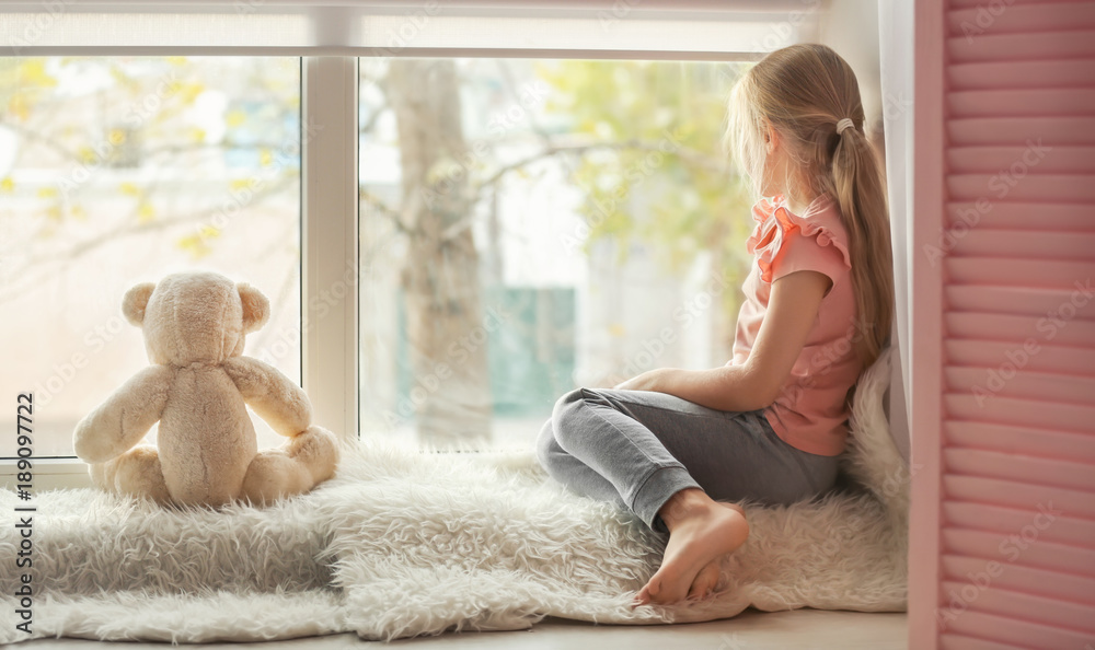 Little girl with teddy bear sitting on window sill. Autism concept